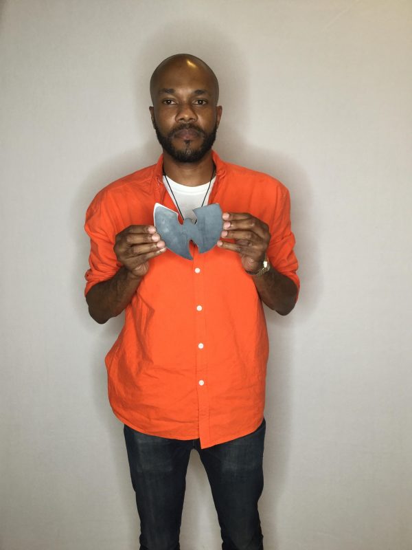 A male-presenting person with brown skin and tightly trimmed brown hair and beard holds up a small object against his bright orange shirt