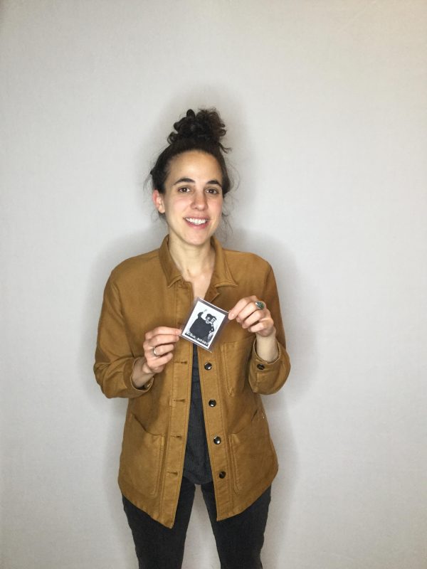 A female-presenting person with curly brown hair pulled up in a bun holds two edges of a small piece of paper in their hands