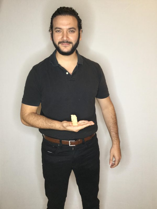 A male-presenting person dressed in all black except for a brown belt displays a small object in the palm of their right hand