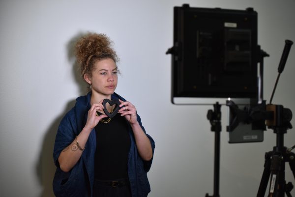 A female person poses with a small heart held close to their chin with both hands in front of a studio light