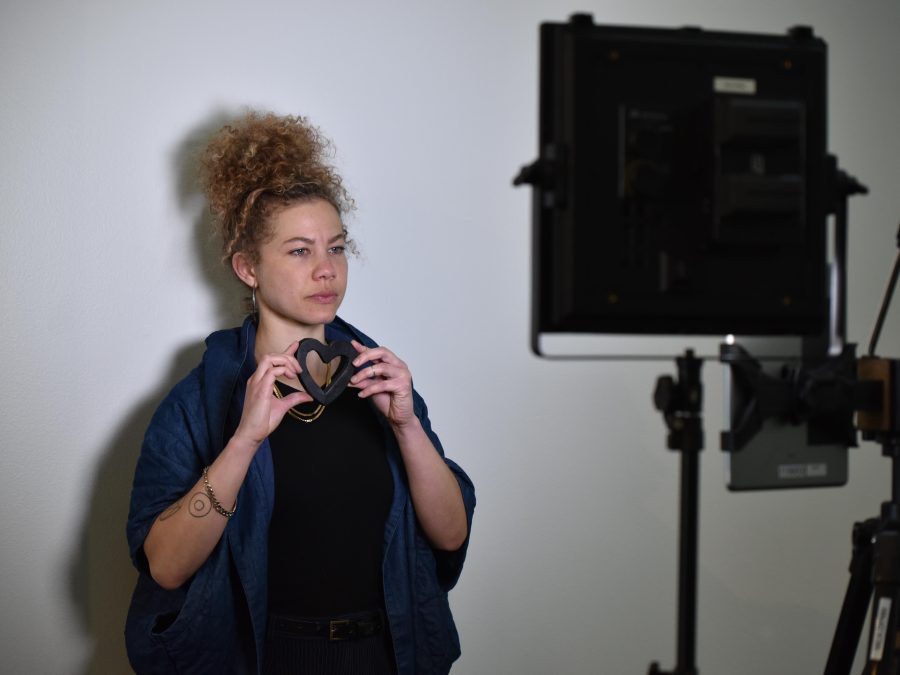 A female person poses with a small heart held close to their chin with both hands in front of a studio light