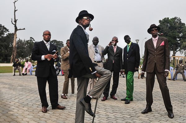 A group of men with various tones of brown skin pose outdoors with suits on.