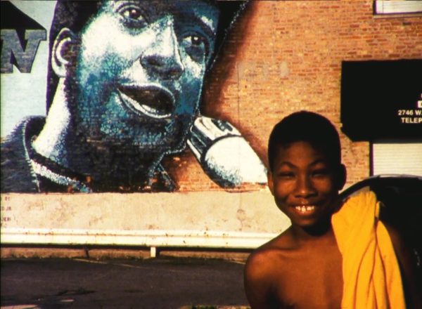 Grainy video still of a young Black person smiling near a mural of Fred Hampton