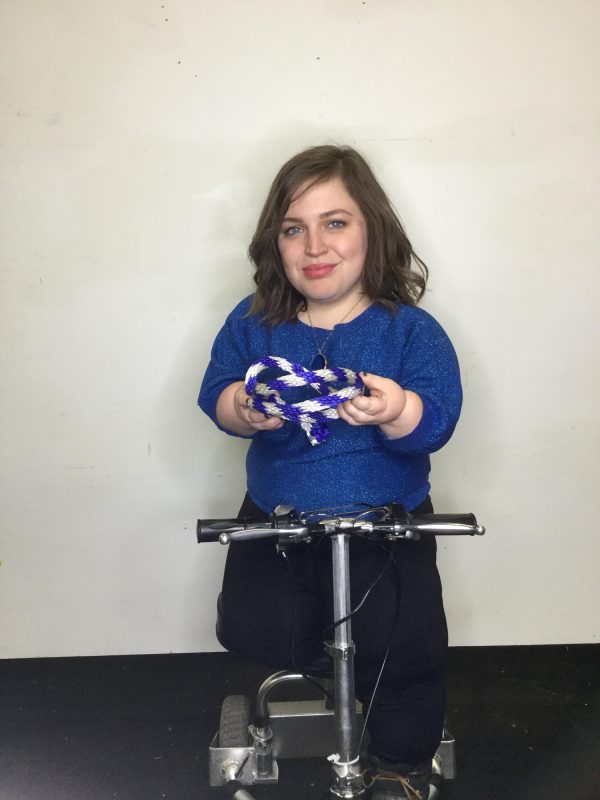 A female-presenting person resting a leg on a scooter holds a purple and white rope in both hands