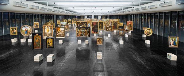 Large open room filled with artworks hanging in rows from the ceiling and white blocks underneath each of them