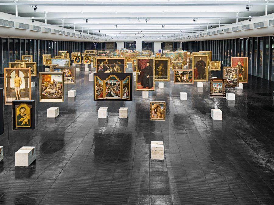 Large open room filled with artworks hanging in rows from the ceiling and white blocks underneath each of them
