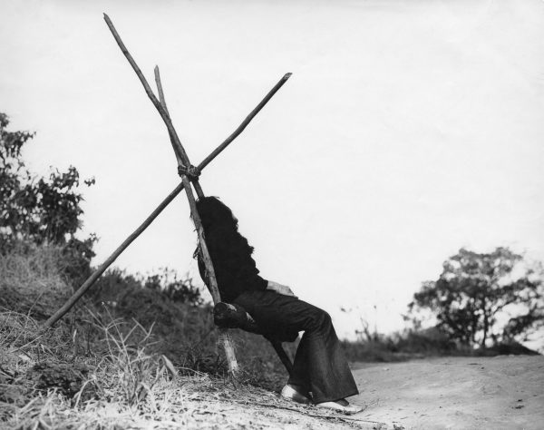 Black and white image of a person sitting on a structure made of tree branches and rope