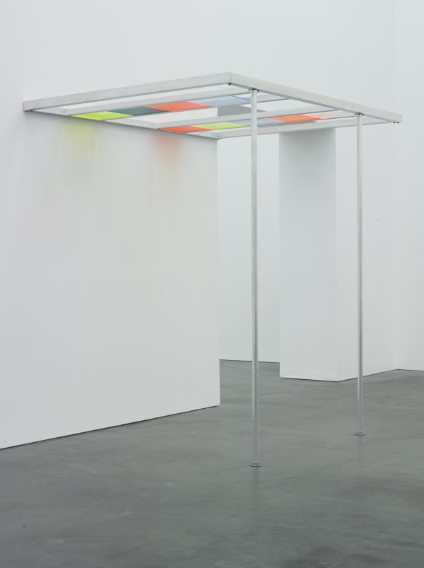 A pergola-like structure held up by two thin metal poles in a white-walled room. The roof of the structure is divided into four rows, two of which have various colored light filters