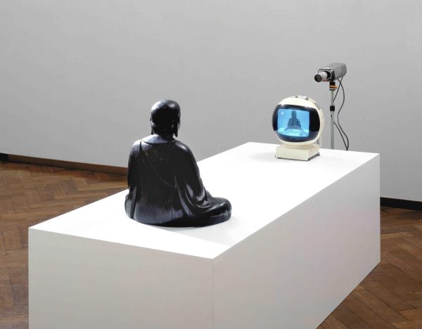 A statue on a white pedestal in front of a tv and camera displaying a live feed of the statue