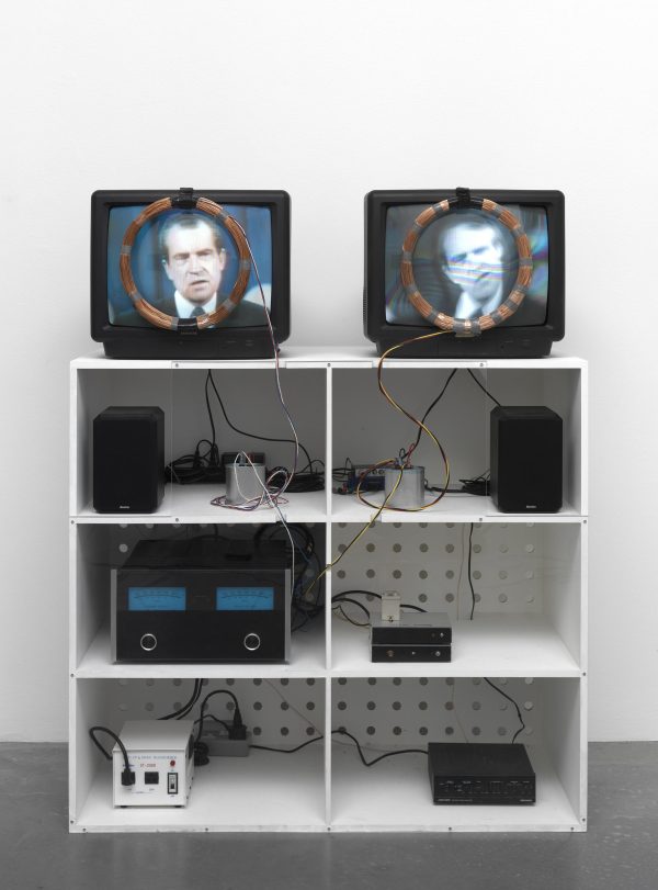 A white shelving unit with various electronics on the shelves. On top are two tvs displaying a still of President Nixon with a wire coil wreath hanging from each to frame the images.