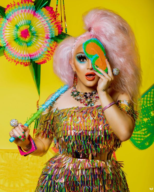 A person with big pink hair, dressed in colorful apparel, jewelry, and makeup, covers their left eye with a mask, with an expression of surprise.
