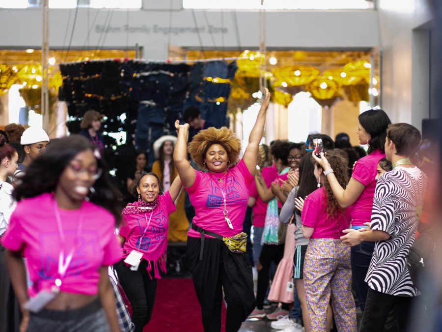 Young people in bright pink t-shirts walk jubilantly down a red carpet as onlookers watch and applaud.