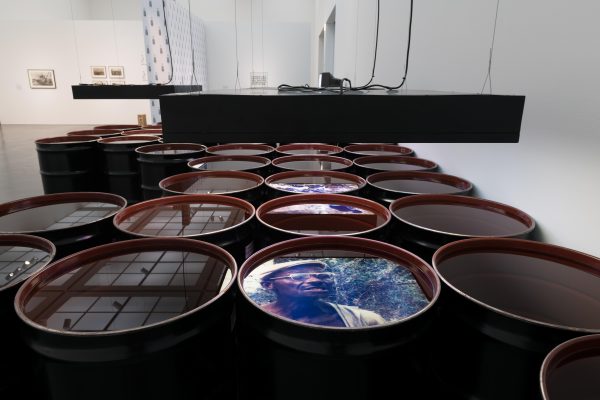 Rows of barrels filled with liquid in a few images are reflected onto the surface. At the forefront is a male presenting person with dark skin against greenery.