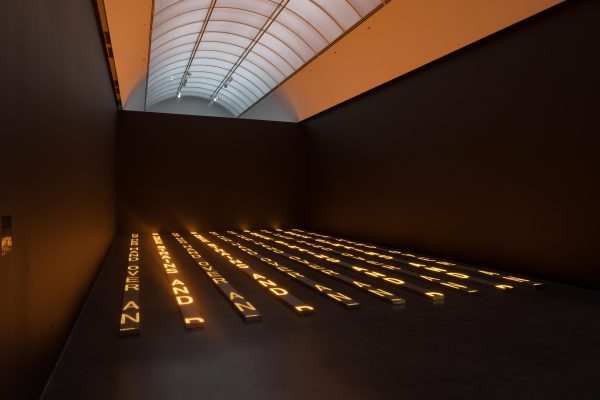 In a large darkened room, large letters are illuminated in yellow in vertical rows on the floor. These letters cast a warm glow over the room.