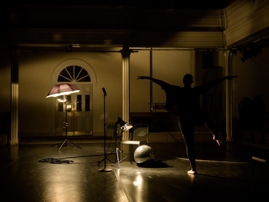 In a dramatically lit setting, a dancer in silhouette stands on one leg in arabesque with their arms extended like wings.
