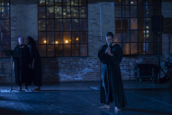 In a dark setting, a person in a long dark robe stands, holding a microphone to their mouth while two other people, similarly dressed, stand together in the background.