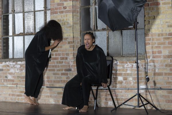 A person sits in a chair against a brick wall with eyes squeezed shut and mouth open, as another person turns towards the brick wall, their face covered with hair.