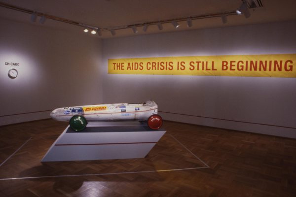 A sleek soap-box derby car on a pedestal in a dimly lit gallery space in front of an illuminated sign that reads The AIDS crisis is still beginning