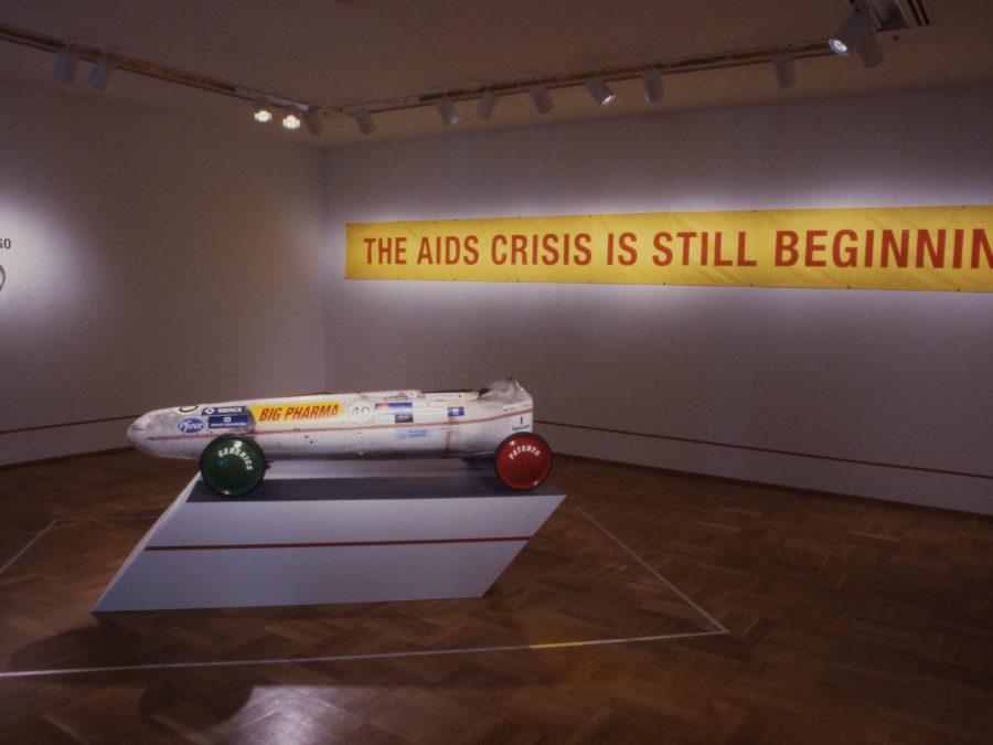 A sleek soap-box derby car on a pedestal in a dimly lit gallery space in front of an illuminated sign that reads The AIDS crisis is still beginning