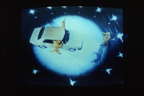 Grainy image of three people in yellow suits surrounding a white sedan in the middle of a spotlight. The image has been captured from above, making it appear as if they are on a planet in space.