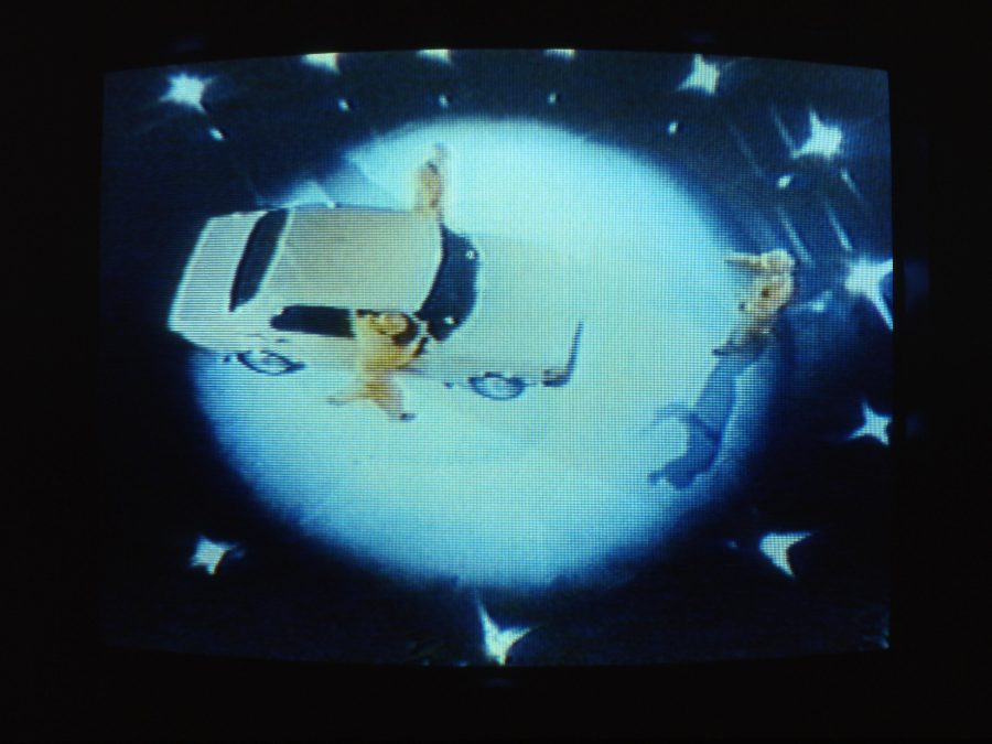 Grainy image of three people in yellow suits surrounding a white sedan in the middle of a spotlight. The image has been captured from above, making it appear as if they are on a planet in space.