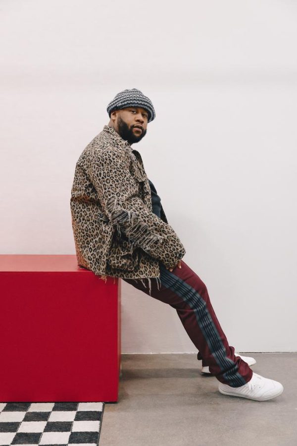 A person with light brown skin and a dark full beard dressed in a animal print coat and red pants with black striped trim on the side sits on a red block