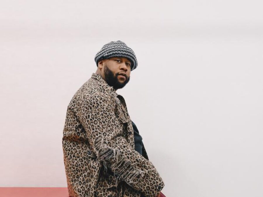 A person with light brown skin and a dark full beard dressed in a animal print coat and red pants with black striped trim on the side sits on a red block