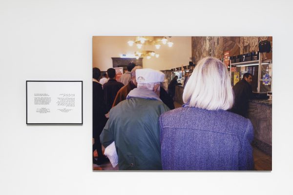 A photograph of two people's heads waiting in a long line for a counter next two a framed text