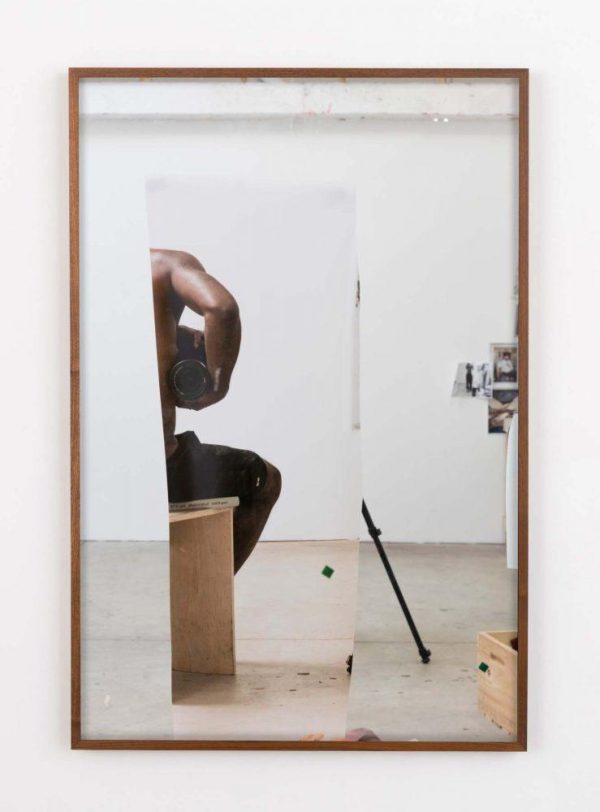 A mirrored reflection shows the right half of a dark-skinned person holding a camera aimed at the mirror under their arm in a stark white room.