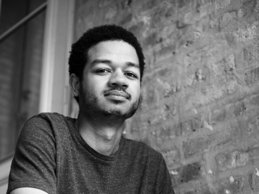 Black and white portrait of a male-presenting person in a short sleeve shirt sitting on set of stairs near a brick wall