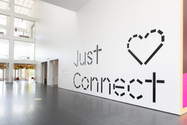 An empty MCA atrium with large black text on one white wall reading Just Connect with a heart emoji stylized in the signature MCA dash font