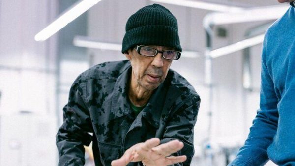 A person in a black collared shirt, black knit hat, and glasses reaches toward something in a brightly lit studio space