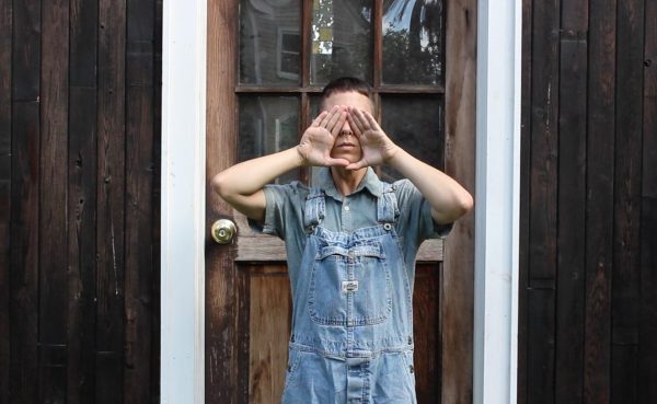 A person in overalls stands next to a wooden door with their hands making a diamond shape over their face to cover their eyes, showing only their nose and mouth