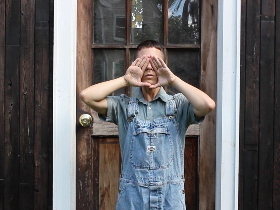 A person in overalls stands next to a wooden door with their hands making a diamond shape over their face to cover their eyes, showing only their nose and mouth