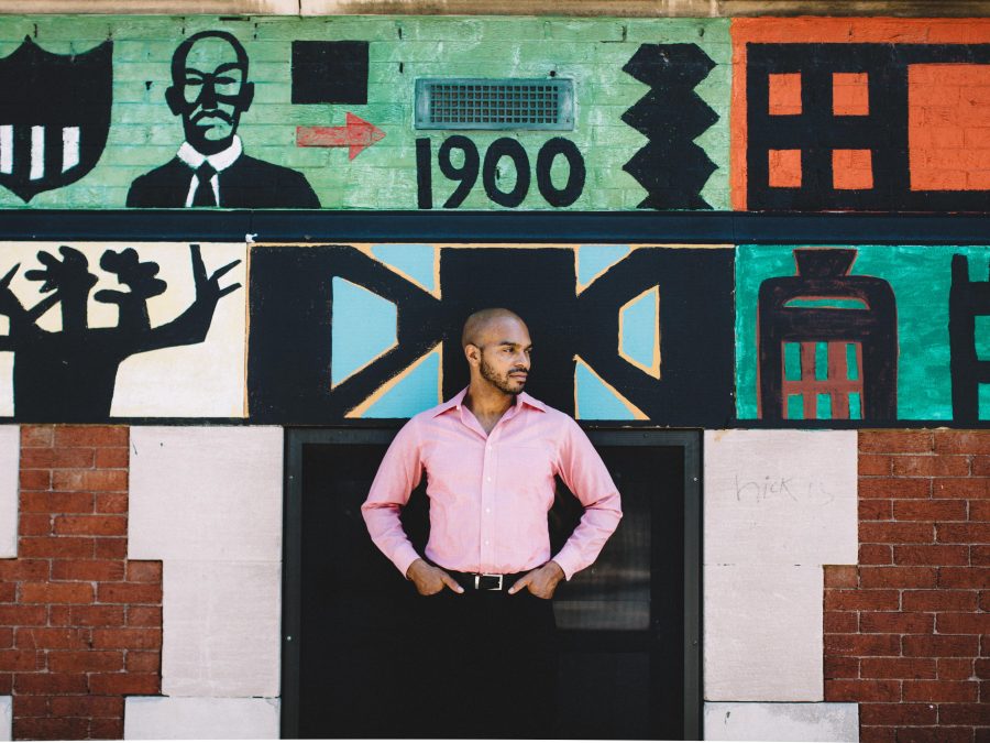 A male-presenting person with medium-brown skin in a pink dress shirt and black paints stands with arms at their hips in front of a colorful mural with the numbers 1900 above their head