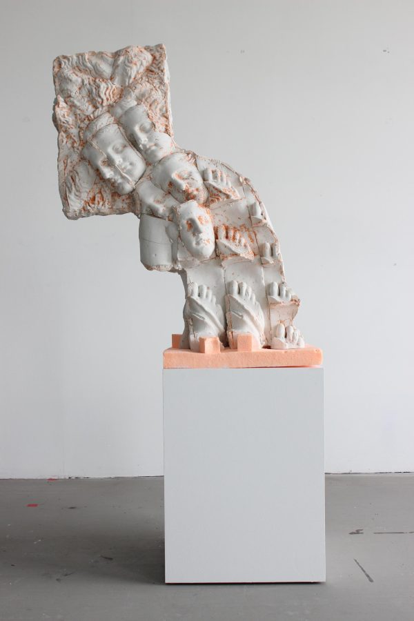 A wave-like sculpture made up of repeating heads and feet on a white pedestal in a gallery space