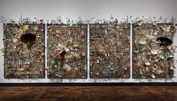 Four ornate panels hang on gallery wall each heavily sculptured with various materials, including ceramic birds, metal flowers, afghans, strung beads, crystals, and an antique gramophone