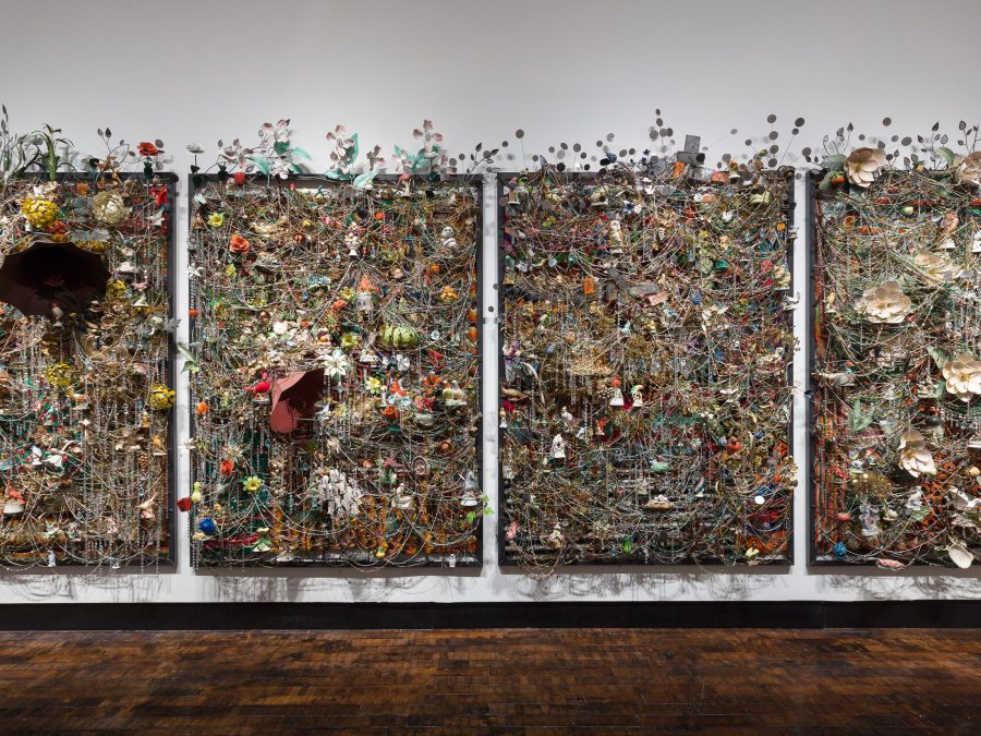 Four ornate panels hang on gallery wall each heavily sculptured with various materials, including ceramic birds, metal flowers, afghans, strung beads, crystals, and an antique gramophone