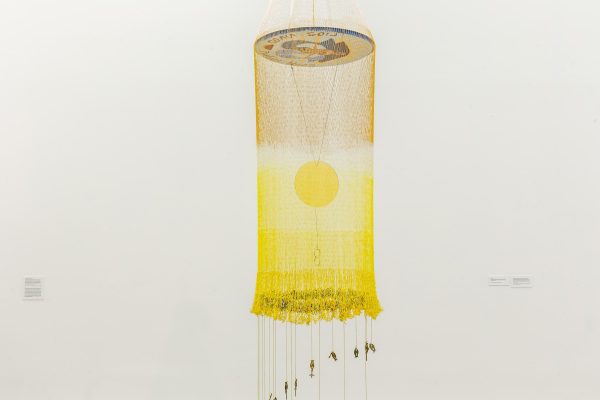 A yellow cylinder made out of strings hangs in a white space