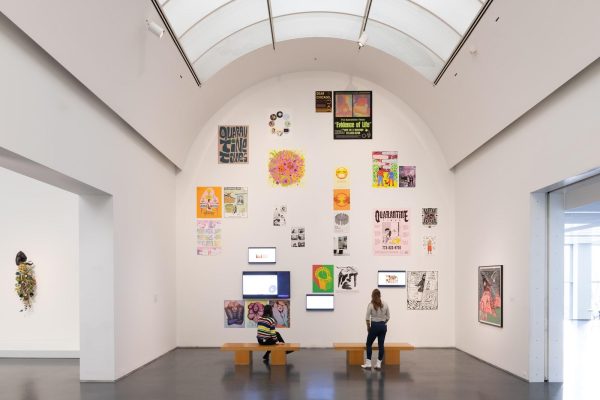 Two people view artworks displayed all over a tall arched white wall in a gallery space