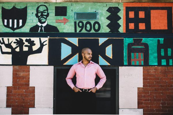 A male-presenting person with medium-brown skin in a pink dress shirt and black paints stands with arms at their hips in front of a colorful mural with the numbers 1900 above their head
