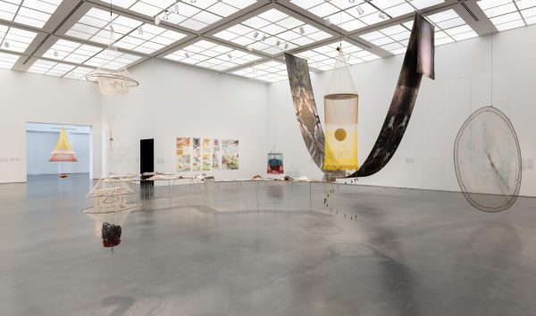 Installation of an exhibition in a white gallery with several artworks on the walls and floor, including a large sheet of fabric draped from the ceiling like a hammock
