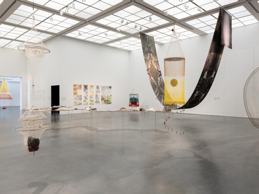 Installation of an exhibition in a white gallery with several artworks on the walls and floor, including a large sheet of fabric draped from the ceiling like a hammock