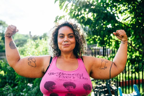 A female presenting person with tanned skin and curly brown hair to their shoulders flexes their biceps in a park. Their pink tank top has text on it that reads Be Beautiful Every Day
