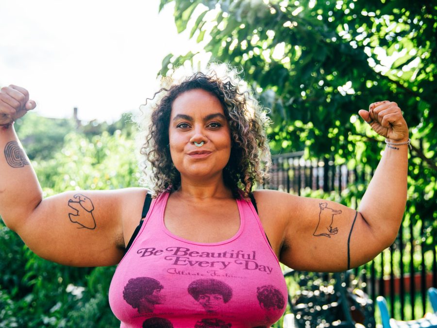 A female presenting person with tanned skin and curly brown hair to their shoulders flexes their biceps in a park. Their pink tank top has text on it that reads Be Beautiful Every Day