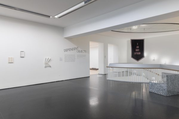 View of a white-walled gallery space near a stairwell. Two openings reveal another room in the distance.