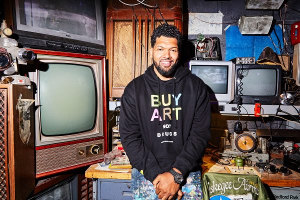 A male-presenting person in a black Buy Art sweatshirt sits on a table while surrounded by different size TVs
