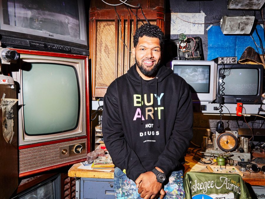 A male-presenting person in a black Buy Art sweatshirt sits on a table while surrounded by different size TVs