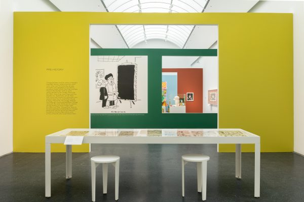 Exhibition interior showing a large white table with in front of a yellow wall with a doorway in the middle