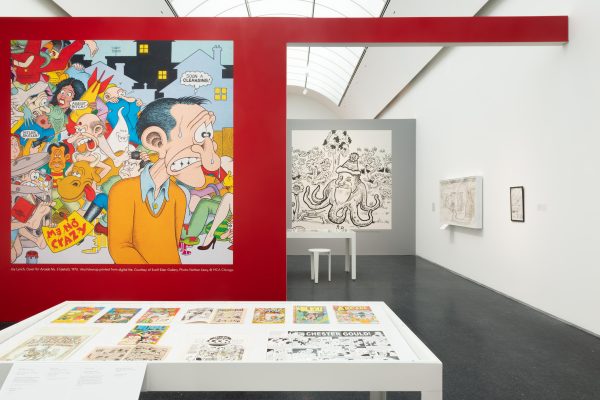 Exhibition interior with a large table and a bold, red wall with comic art depicting a sweating man